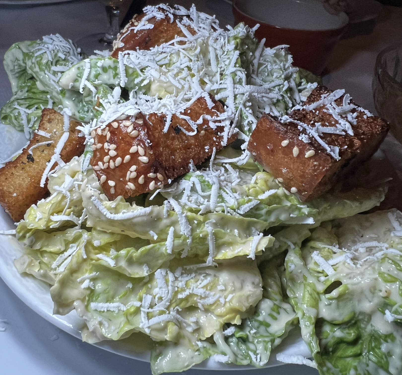 A plate of Caesar salad topped with sesame-crusted croutons and grated cheese, with romaine lettuce coated in creamy dressing.