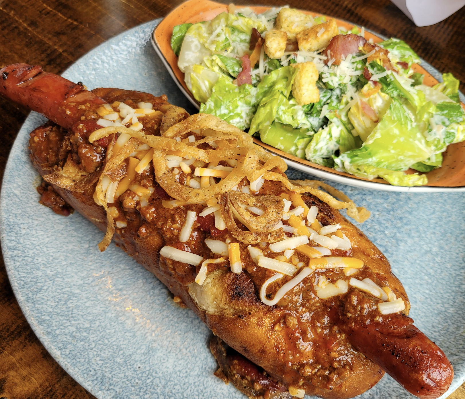 A chili cheese hot dog topped with shredded cheese and fried onions sits on a plate next to a bowl of Caesar salad with croutons and bacon.
