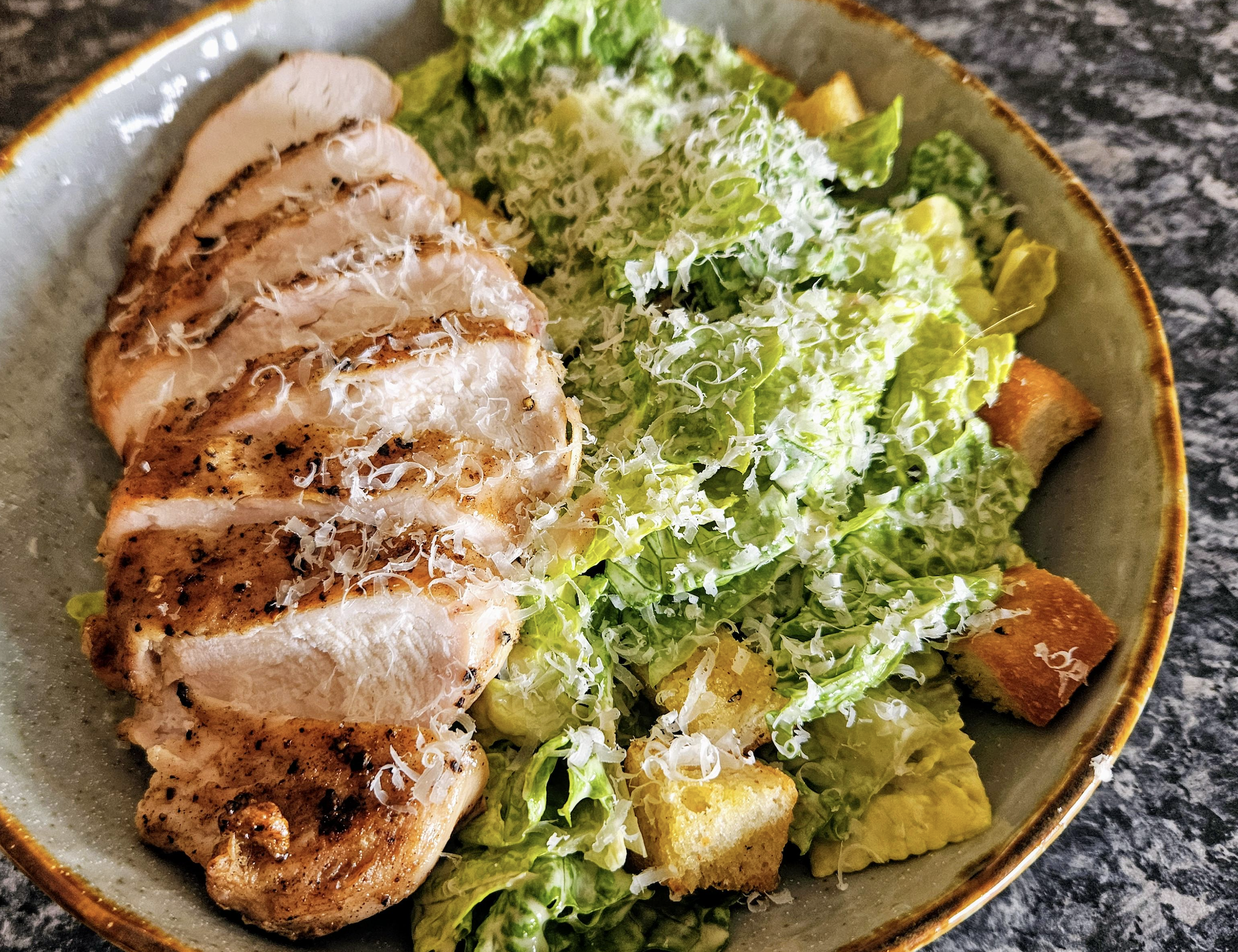 A plate with sliced grilled chicken breast next to Caesar salad topped with grated cheese and croutons.