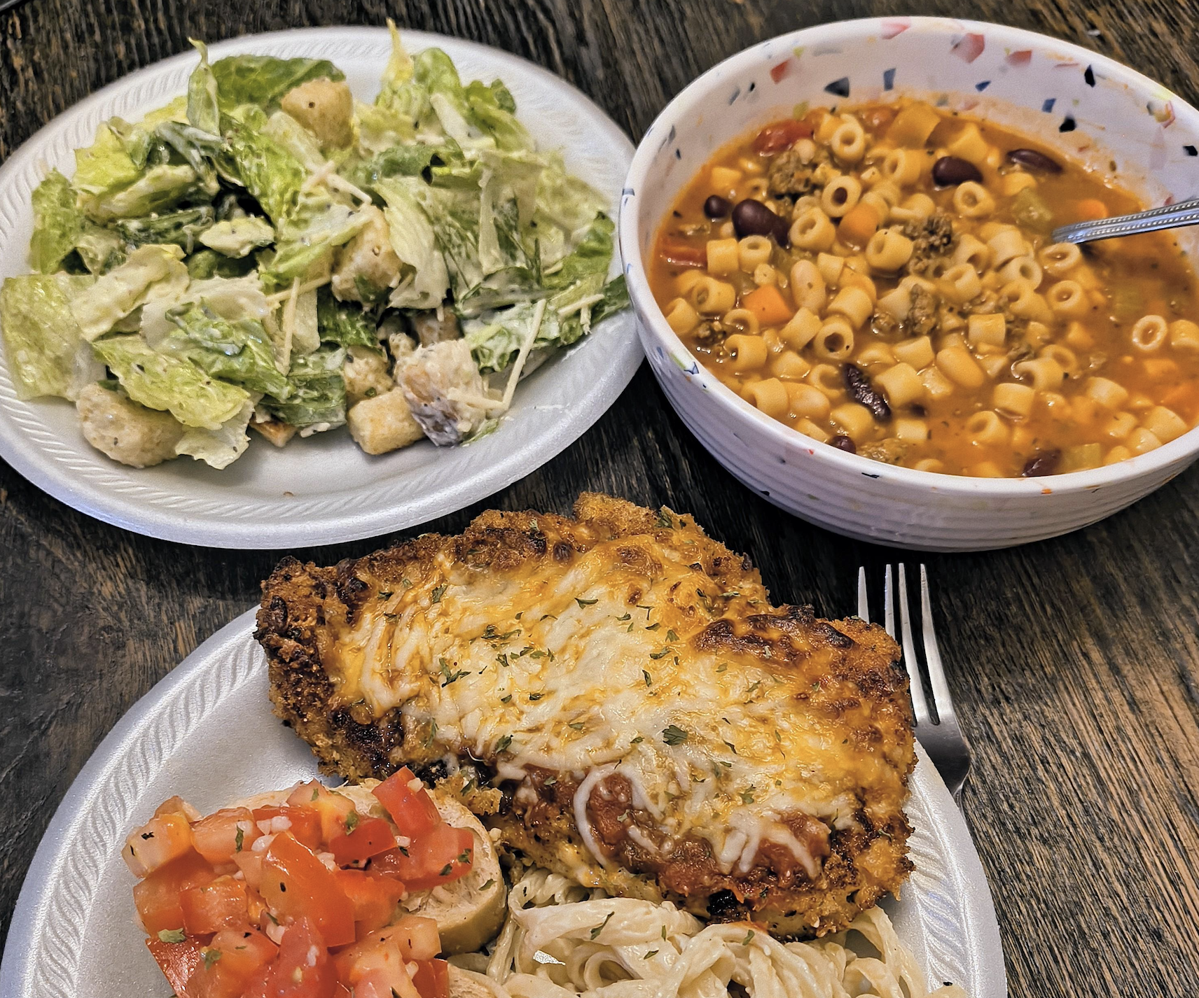 A table with a Caesar salad, a bowl of pasta and bean soup, and a plate with breaded chicken topped with cheese, bruschetta, and pasta in cream sauce.