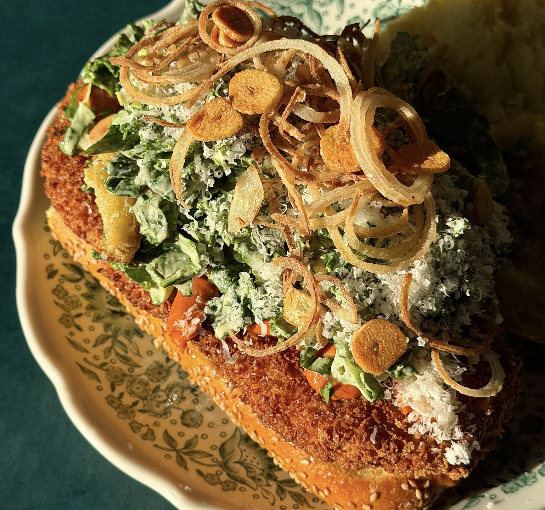 A sandwich on a patterned plate topped with crispy fried onions, fresh greens, and grated cheese, with sunlight highlighting its textures.