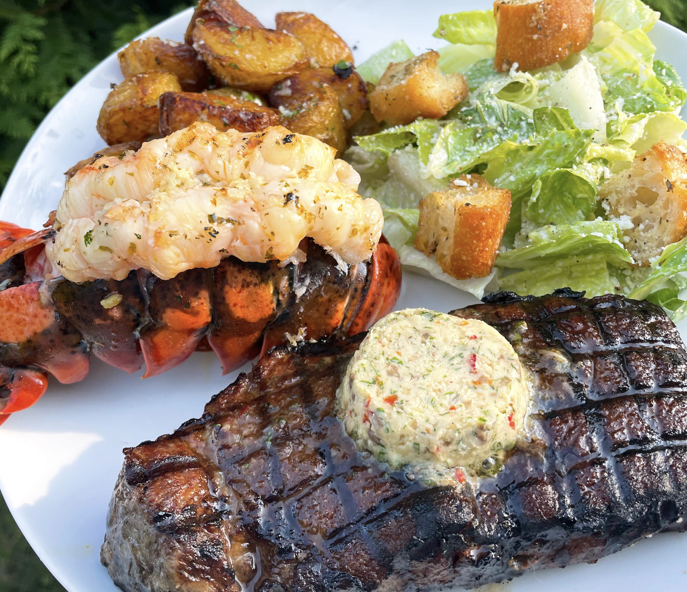 A plate with grilled lobster tail, seasoned potato wedges, Caesar salad with croutons, and a grilled steak topped with herb butter.
