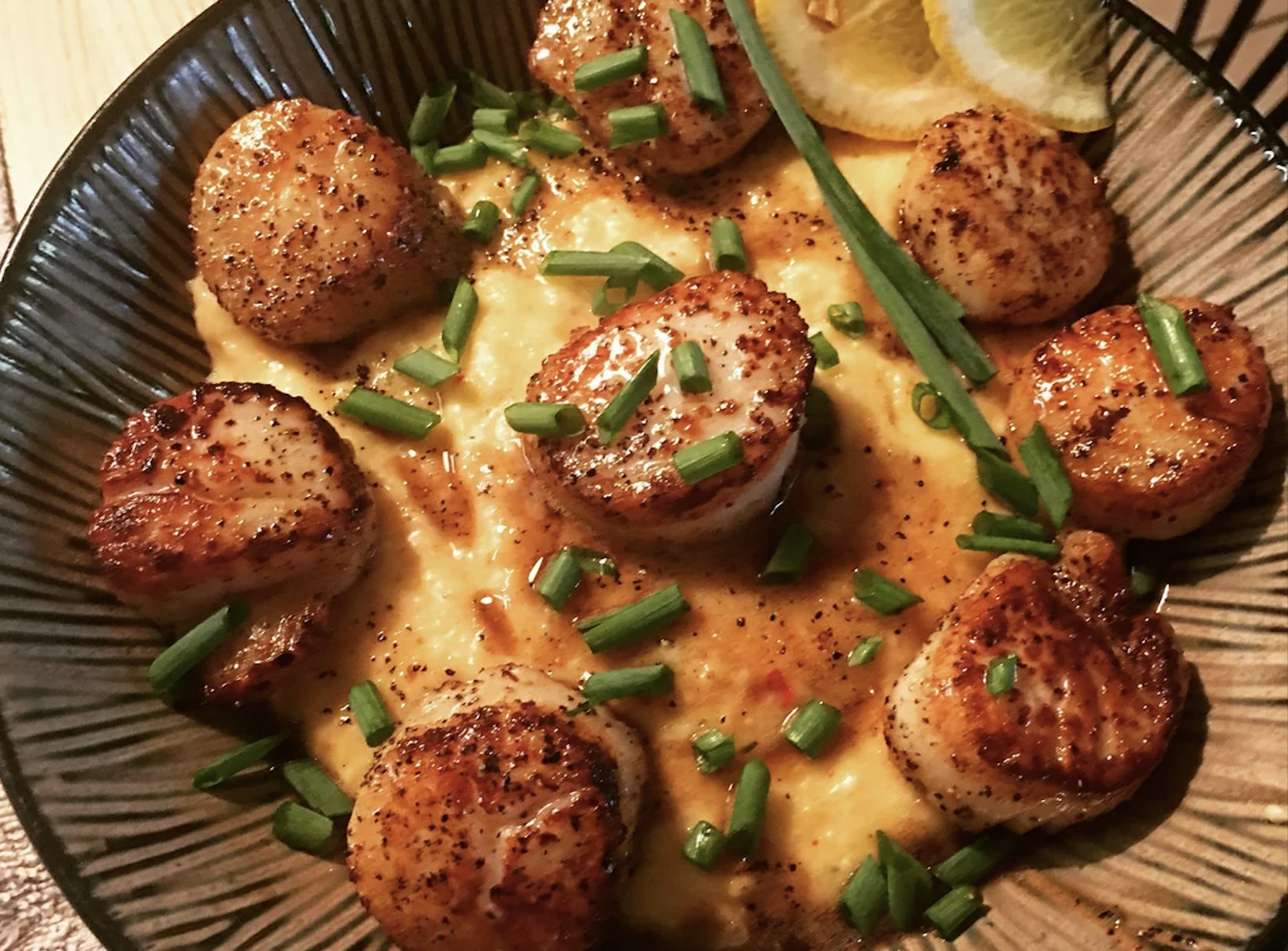 Seared scallops with a peppery crust served over creamy grits, garnished with chopped chives and lemon wedges in a dark bowl.