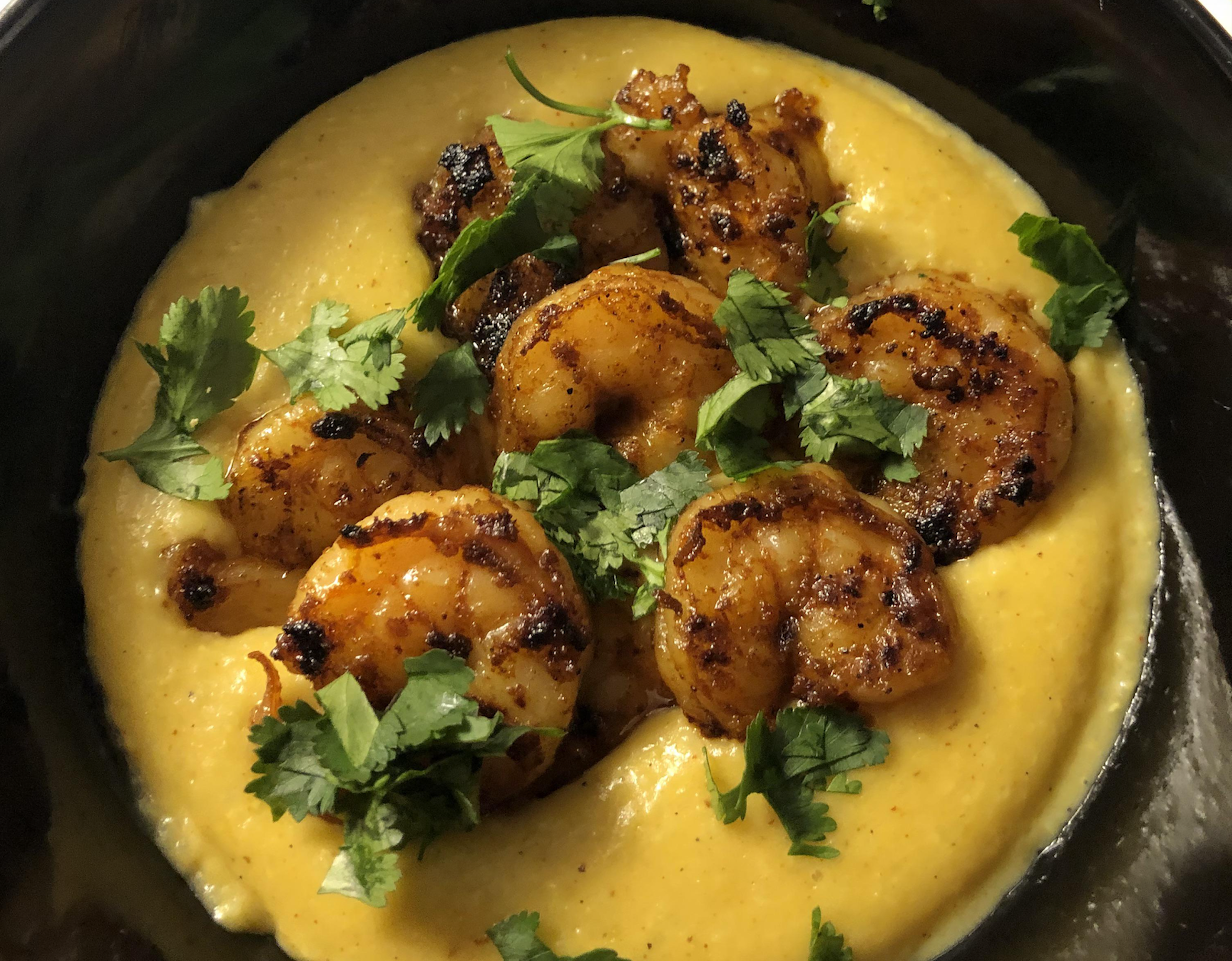 Grilled shrimp coated in spices served on a creamy bed of yellow grits, garnished with fresh cilantro leaves.