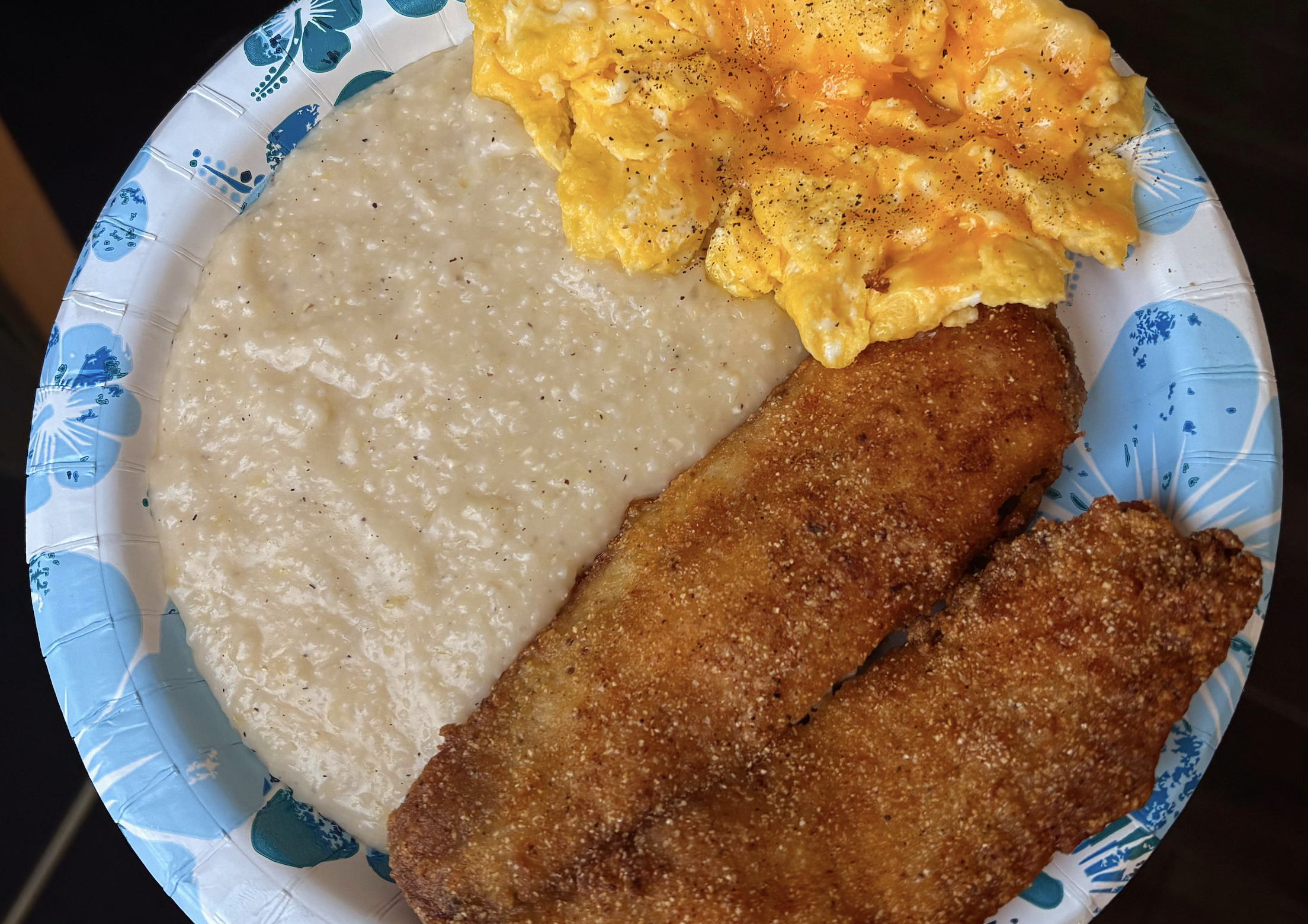 A paper plate with scrambled eggs, creamy grits, and two pieces of fried fish, sprinkled with black pepper.