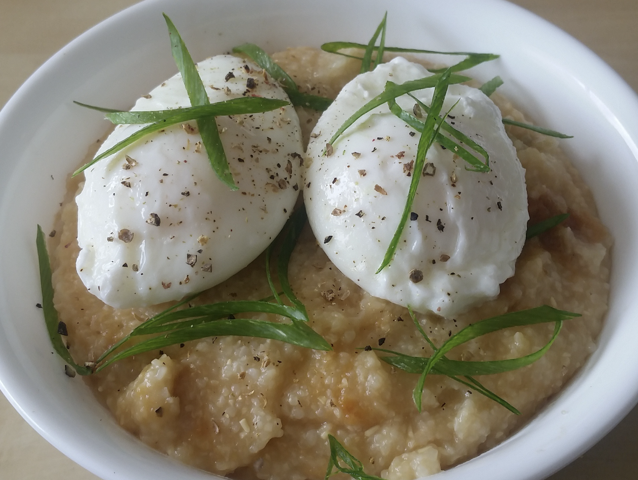 A white bowl of savory oatmeal topped with two poached eggs, sliced green onions, and cracked black pepper.