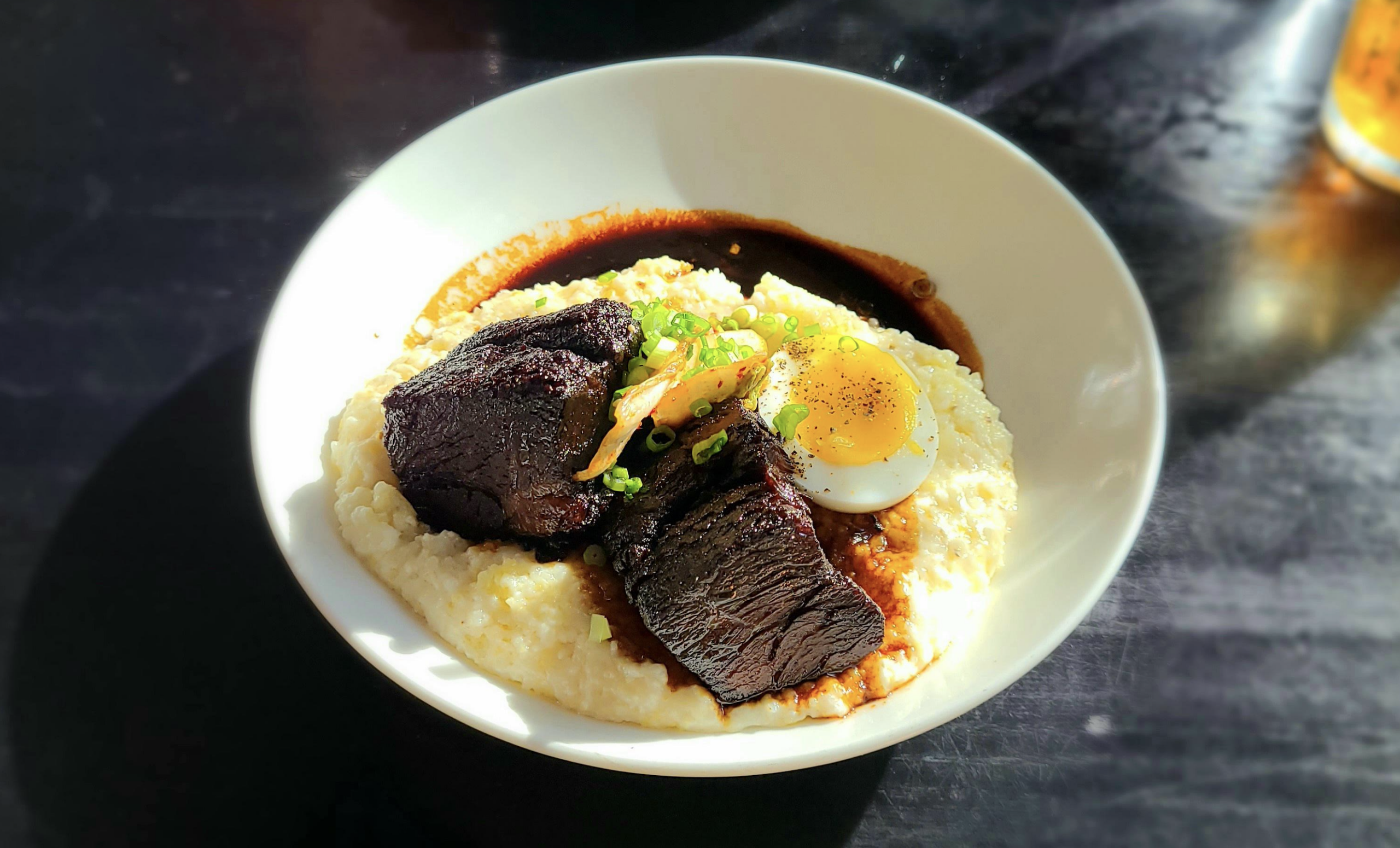 A bowl of creamy grits topped with two pieces of braised short rib, a soft-boiled egg, chopped green onions, pickled vegetables, and a rich brown sauce.