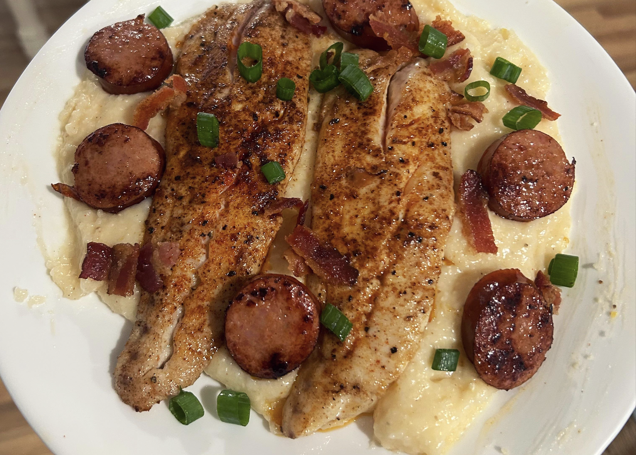 Two seasoned fish fillets on creamy grits, topped with sliced sausage, crispy bacon pieces, and chopped green onions on a white plate.