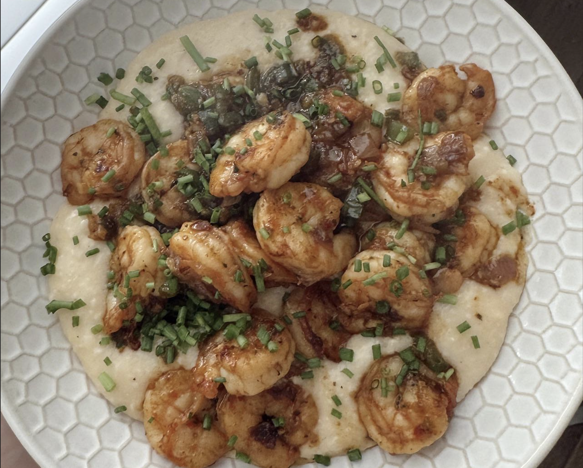 A plate of creamy grits topped with sautéed shrimp, chopped chives, and a savory sauce, served on a hexagon-patterned white dish.