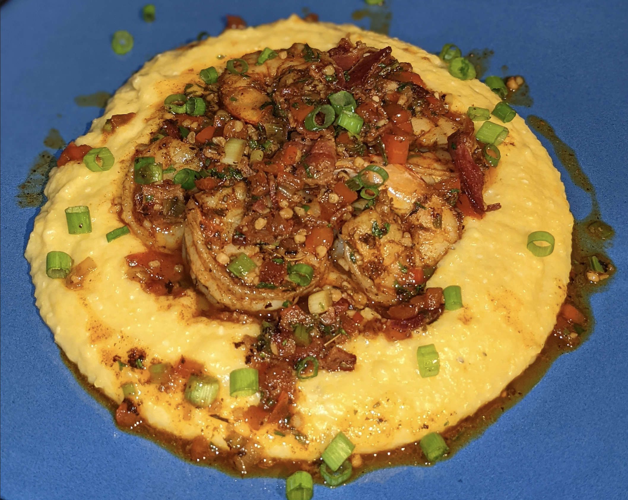 A plate of creamy yellow grits topped with sautéed shrimp, diced vegetables, sauce, and chopped green onions, served on a blue plate.