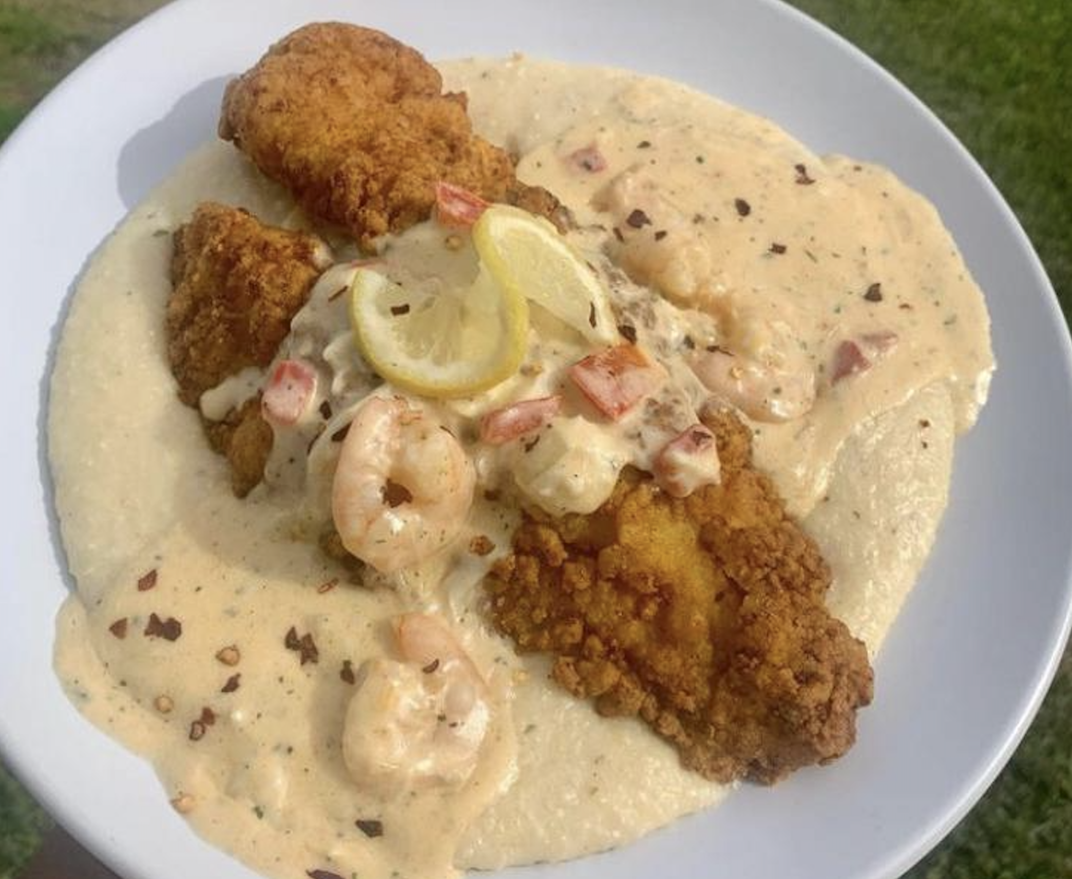 A plate of fried fish fillets over creamy grits, topped with shrimp, diced tomatoes, creamy sauce, and two lemon slices, served outdoors on a white plate.