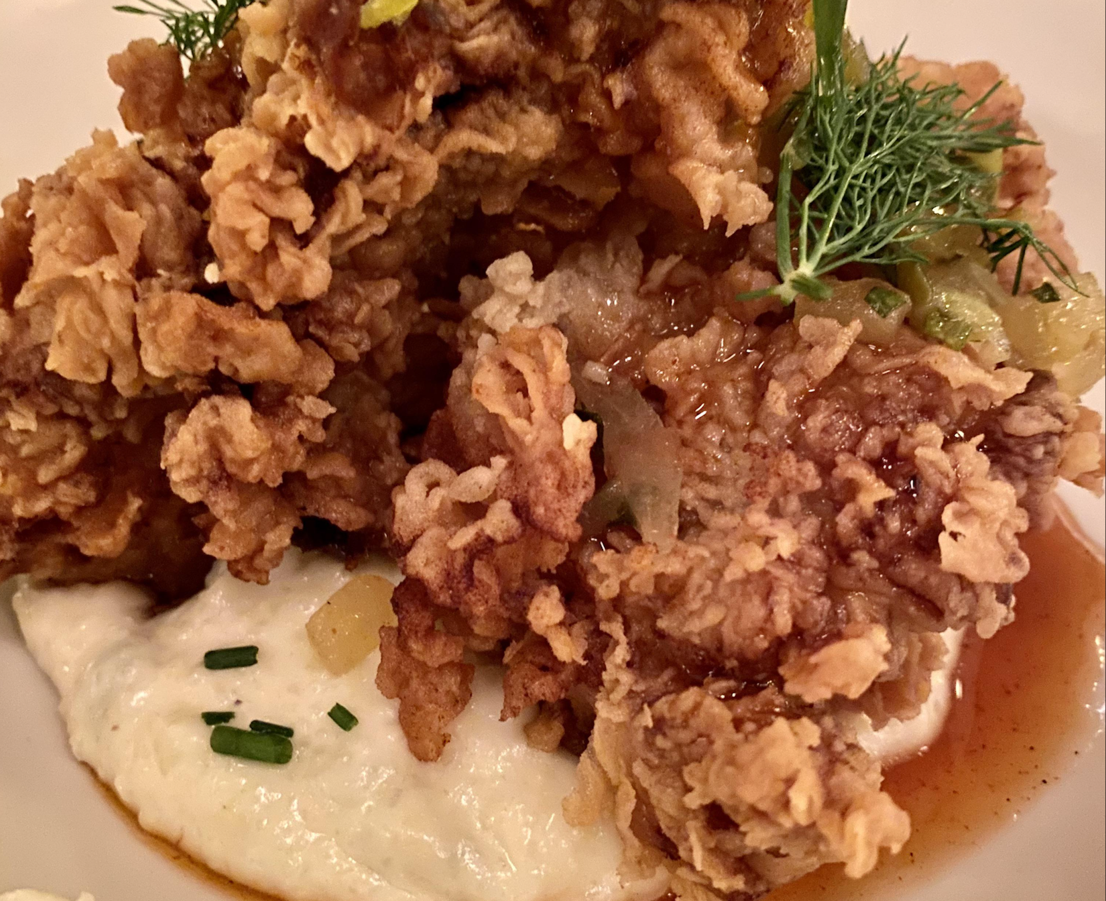 Close-up of crispy fried chicken atop a creamy mashed potato base, garnished with fresh herbs and topped with a drizzle of brown sauce.