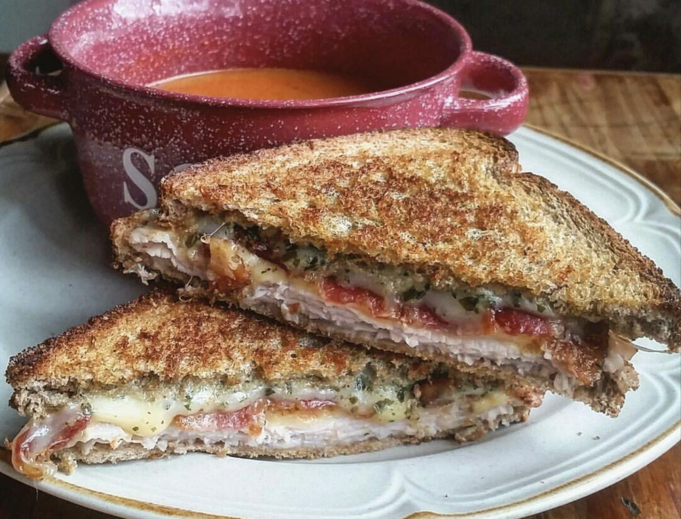 A grilled sandwich with melted cheese and turkey is cut in half and stacked on a plate, with tomato soup in a red bowl behind it.