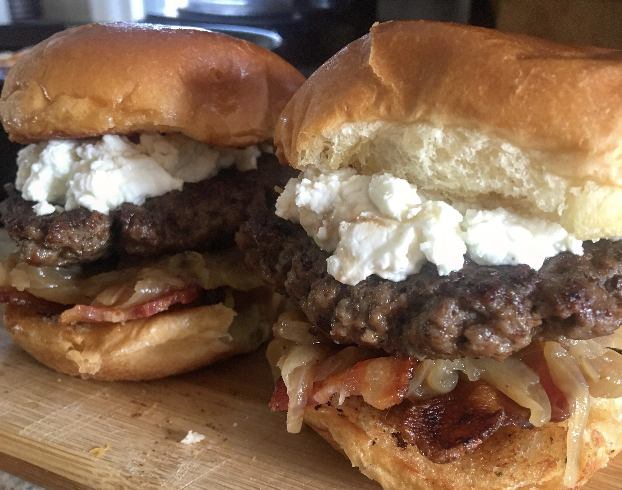 Two beef burgers on toasted buns with caramelized onions, crispy bacon, and a generous topping of crumbled white cheese, placed on a wooden cutting board.