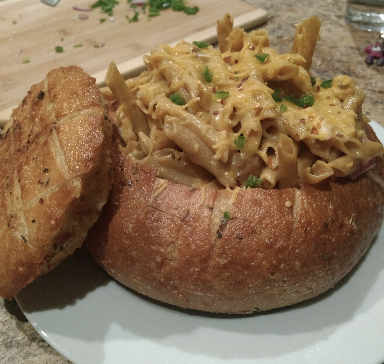 A bread bowl filled with cheesy baked penne pasta sits on a white plate. The bread bowl’s top is placed to the side, and chopped chives garnish the pasta. A cutting board is in the background.