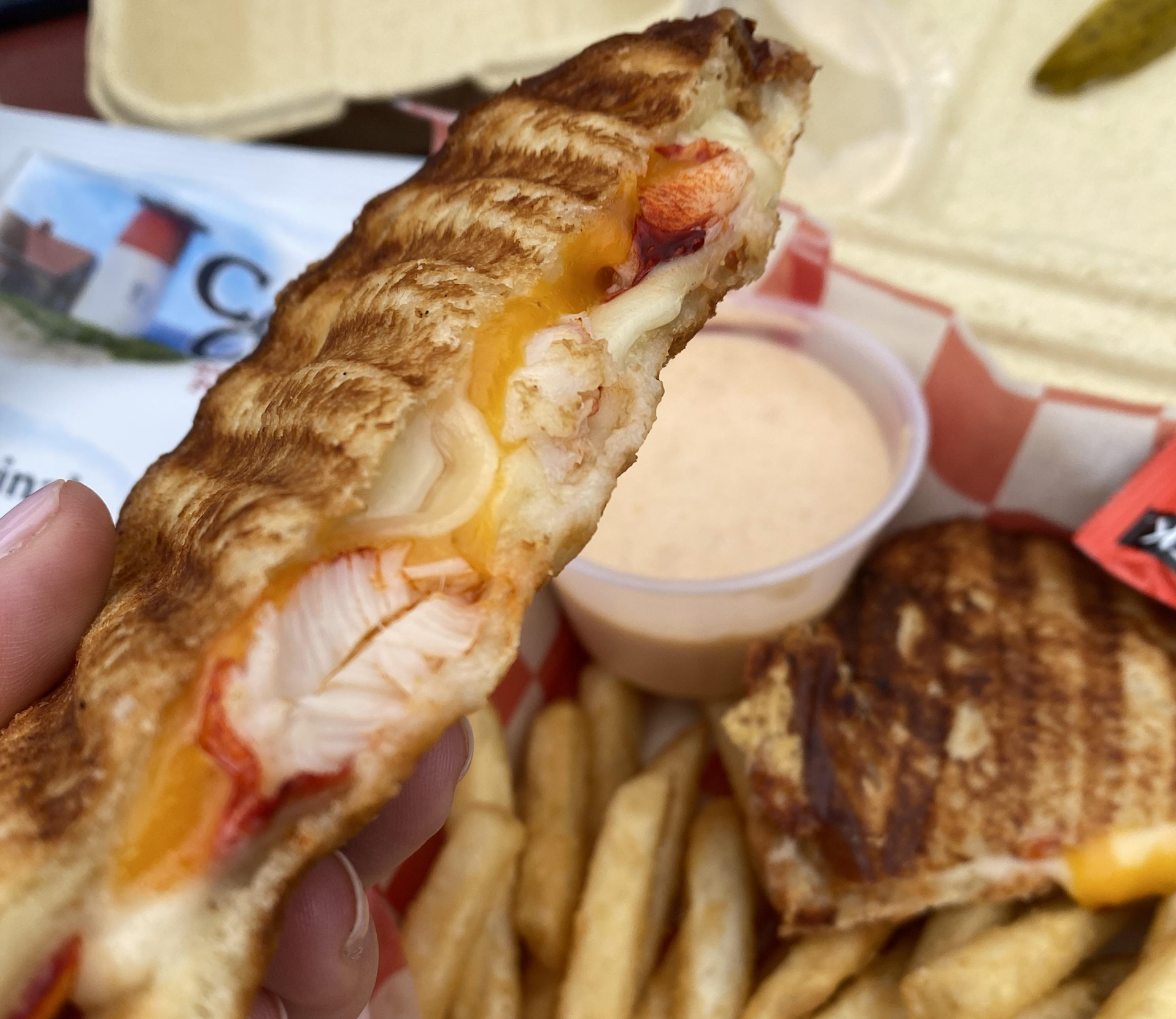 A hand holds a grilled sandwich filled with lobster and melted cheese. French fries, sauce in a cup, and another sandwich half are in the background.