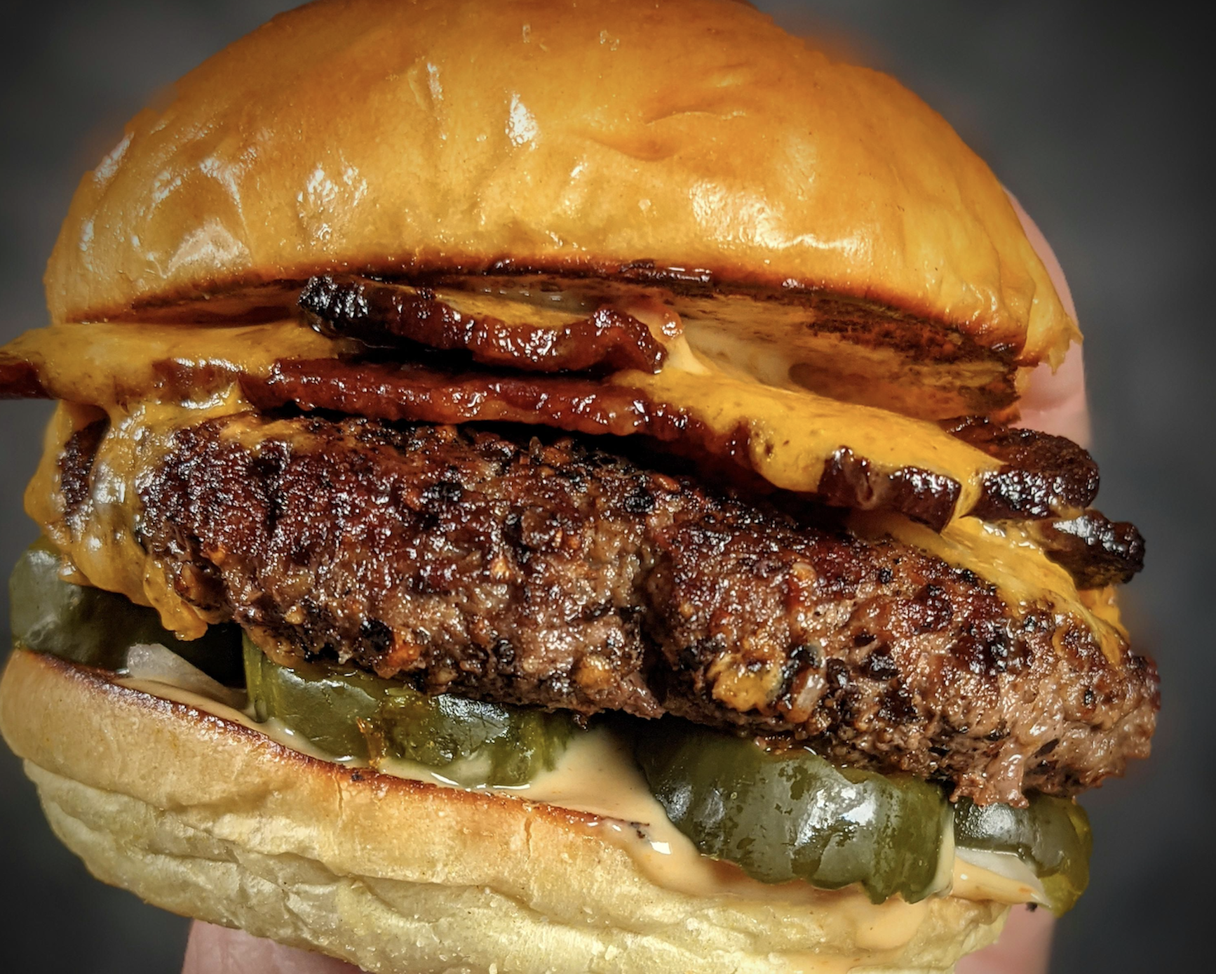 A close-up of a cheeseburger with a beef patty, crispy bacon, melted cheddar cheese, pickles, and sauce in a shiny, toasted bun.