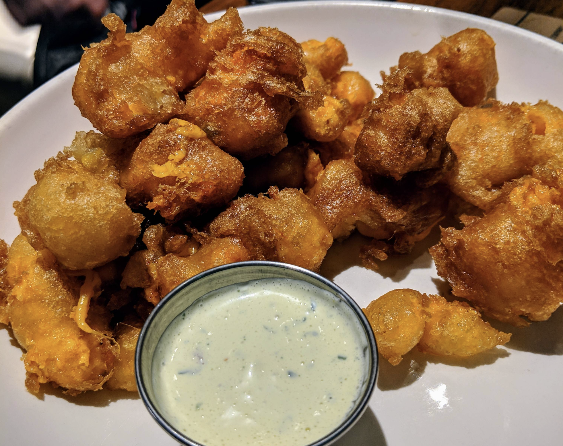 A plate of golden-brown fried cheese curds is served with a small metal cup of creamy dipping sauce.