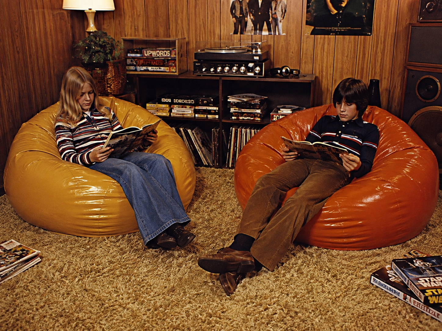 Two kids sit in yellow and orange bean bag chairs reading magazines in a wood-paneled room with records, board games, and a turntable on a shelf, capturing a retro 1970s vibe.