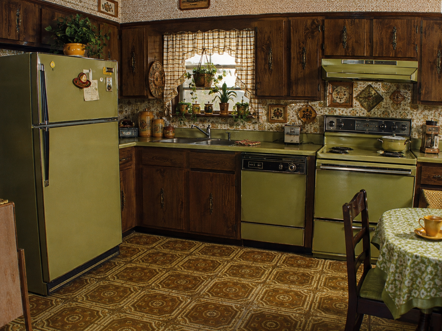 A vintage kitchen with green appliances, dark wood cabinets, patterned wallpaper, and brown linoleum flooring. Plants sit on the windowsill, and a table with a green tablecloth and chairs is in the foreground.