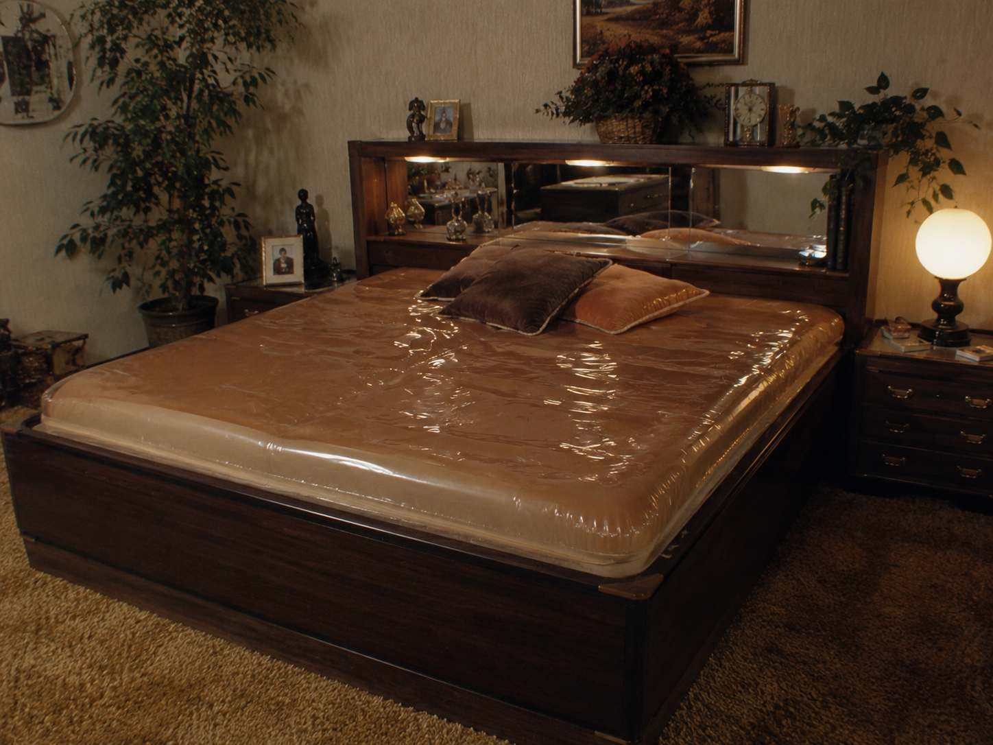 A vintage bedroom with a large waterbed, brown bedspread, pillows, wood headboard with mirror, plants, a glowing lamp, and retro decor.