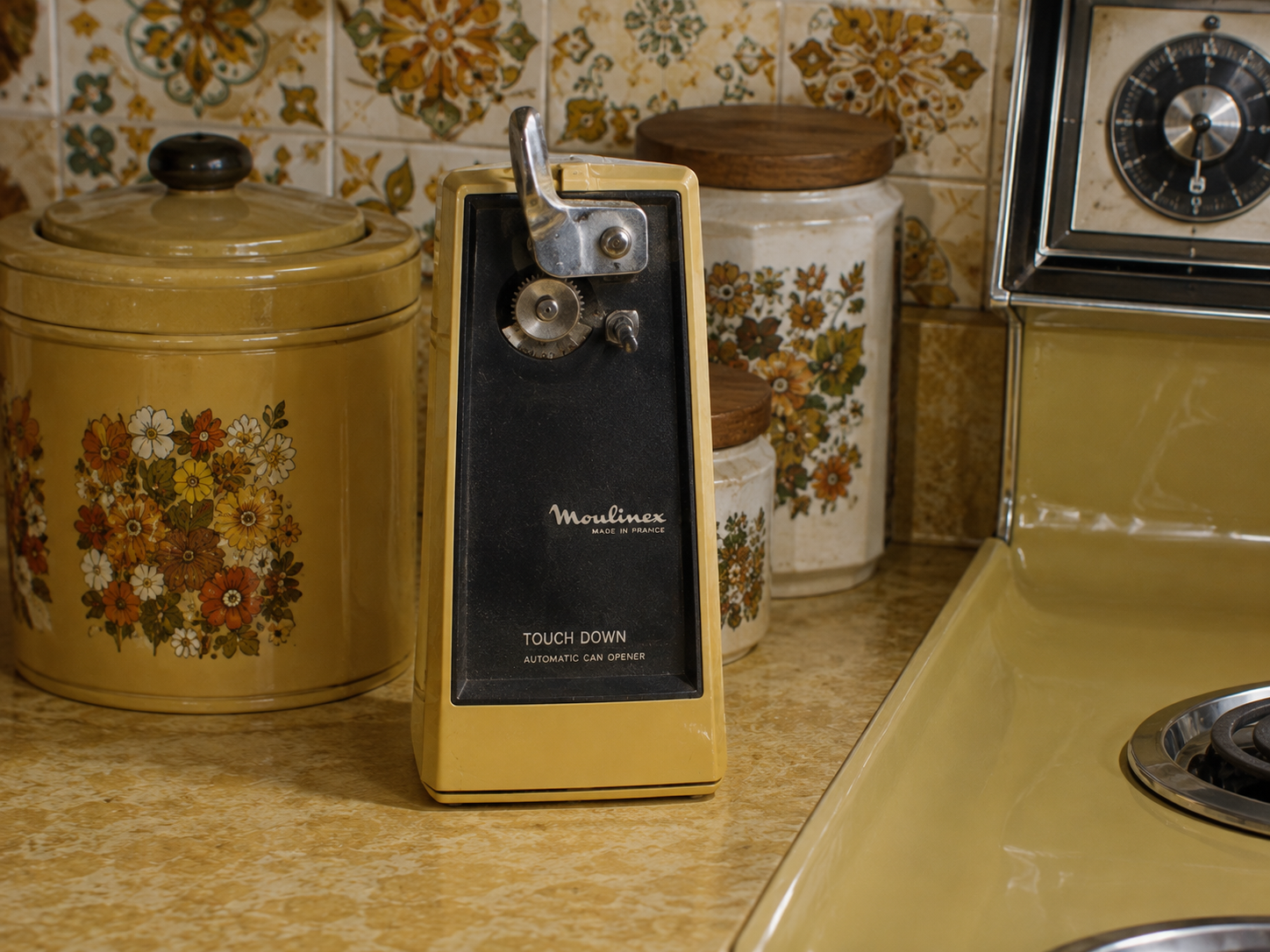 A vintage Moulinex electric can opener sits on a yellow kitchen counter, surrounded by floral-patterned canisters and a retro stove with a clock. The scene has a nostalgic, 1970s feel.