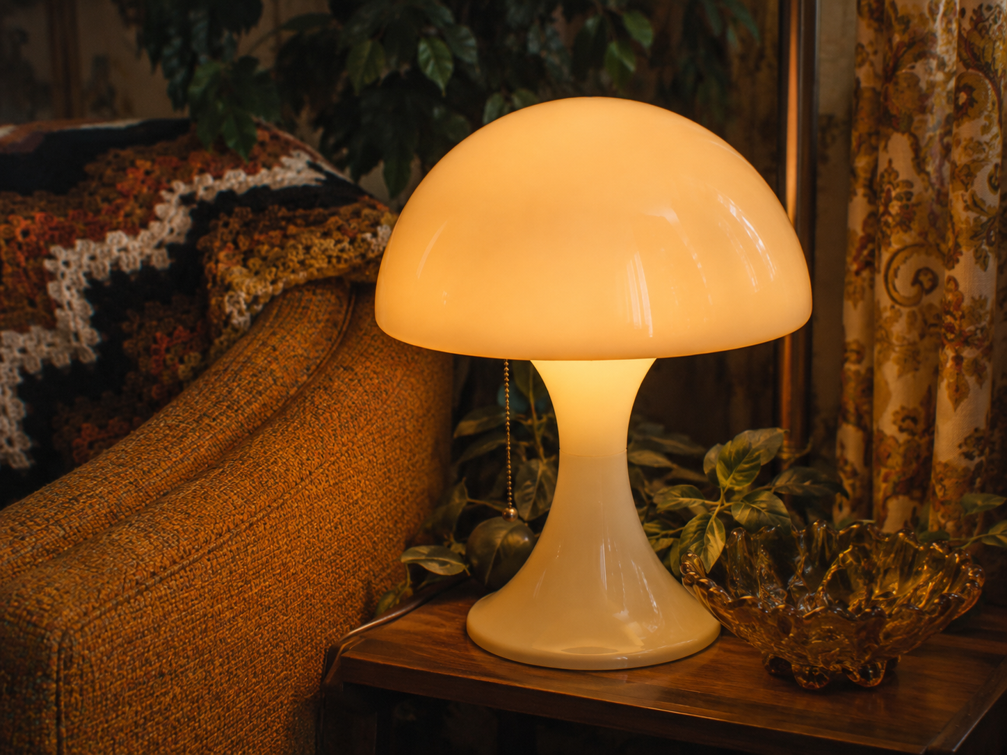 A glowing mushroom-shaped lamp sits on a wooden table next to a golden decorative bowl, with a patterned blanket on an orange couch and green houseplants in the background.