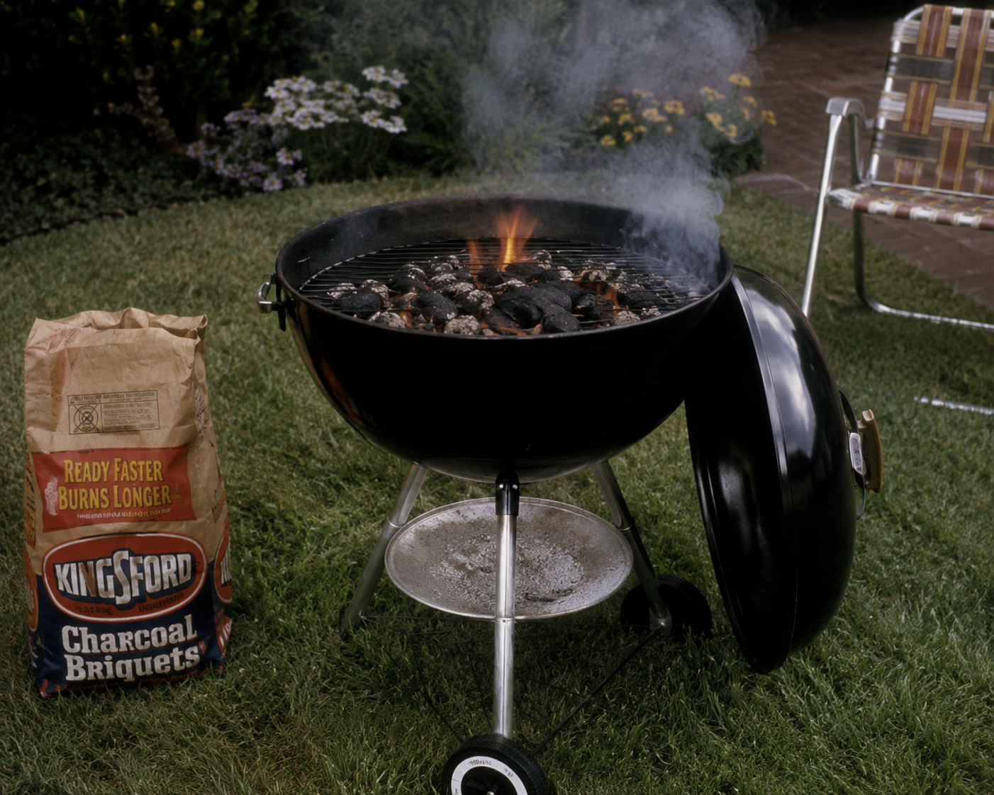 A charcoal grill with flames is on a lawn next to a bag of Kingsford Charcoal Briquets. Smoke rises as the grill lid rests on the ground. A lawn chair and flowers are in the background. 1980s backyard barbecue.