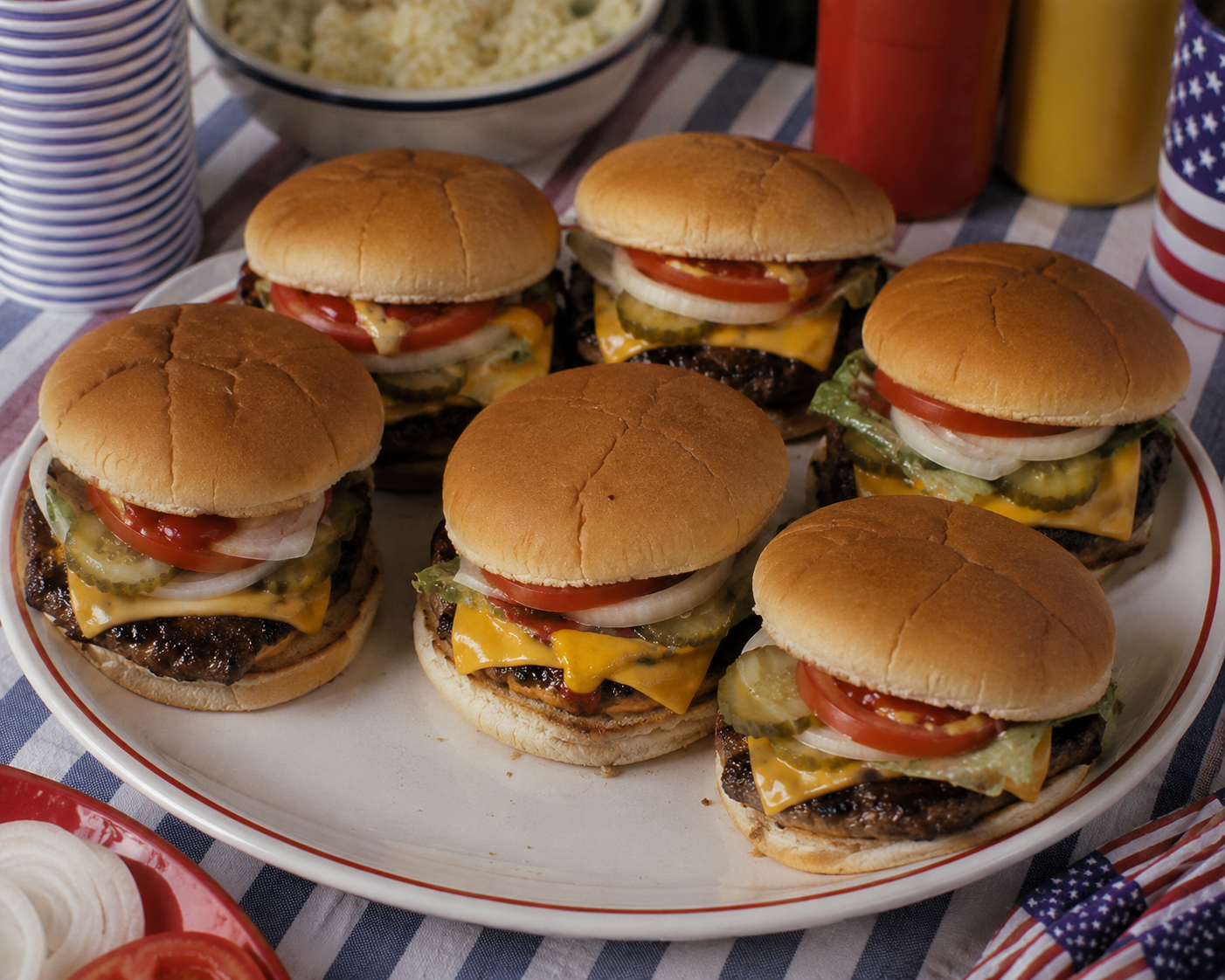 Six cheeseburgers with lettuce, tomato, onion, and pickles sit on a platter. A striped tablecloth, condiments, and American flag napkins are in the background.