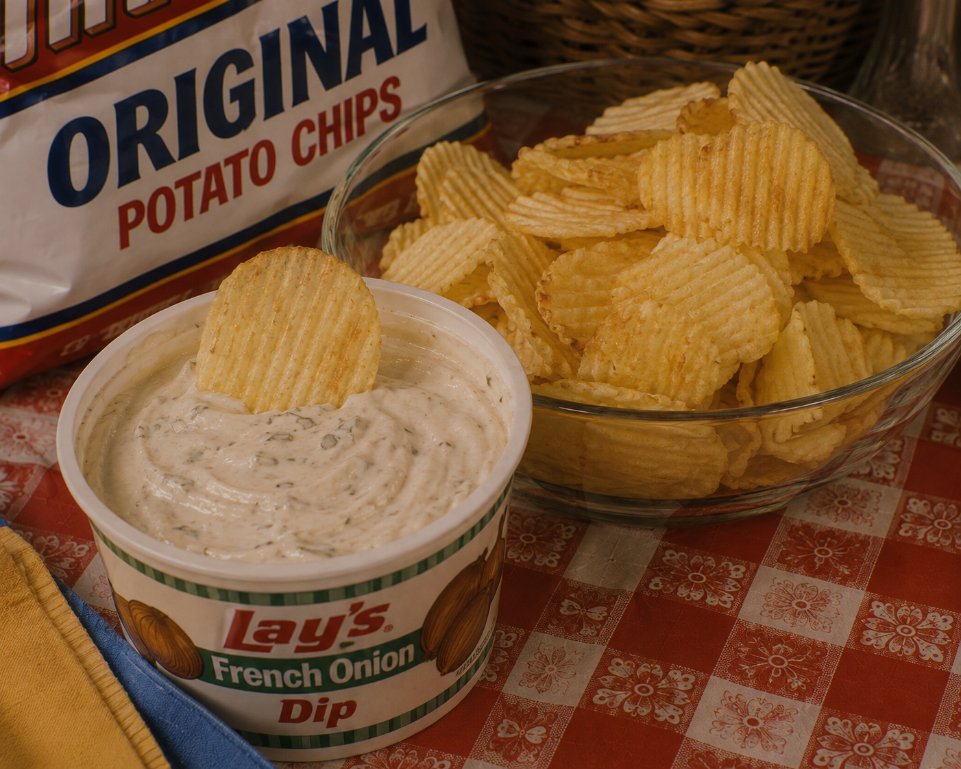A bowl of ridged potato chips sits beside a tub of Lay’s French Onion Dip with one chip dipped. A bag labeled “Original Potato Chips” is in the background on a red-checkered tablecloth.