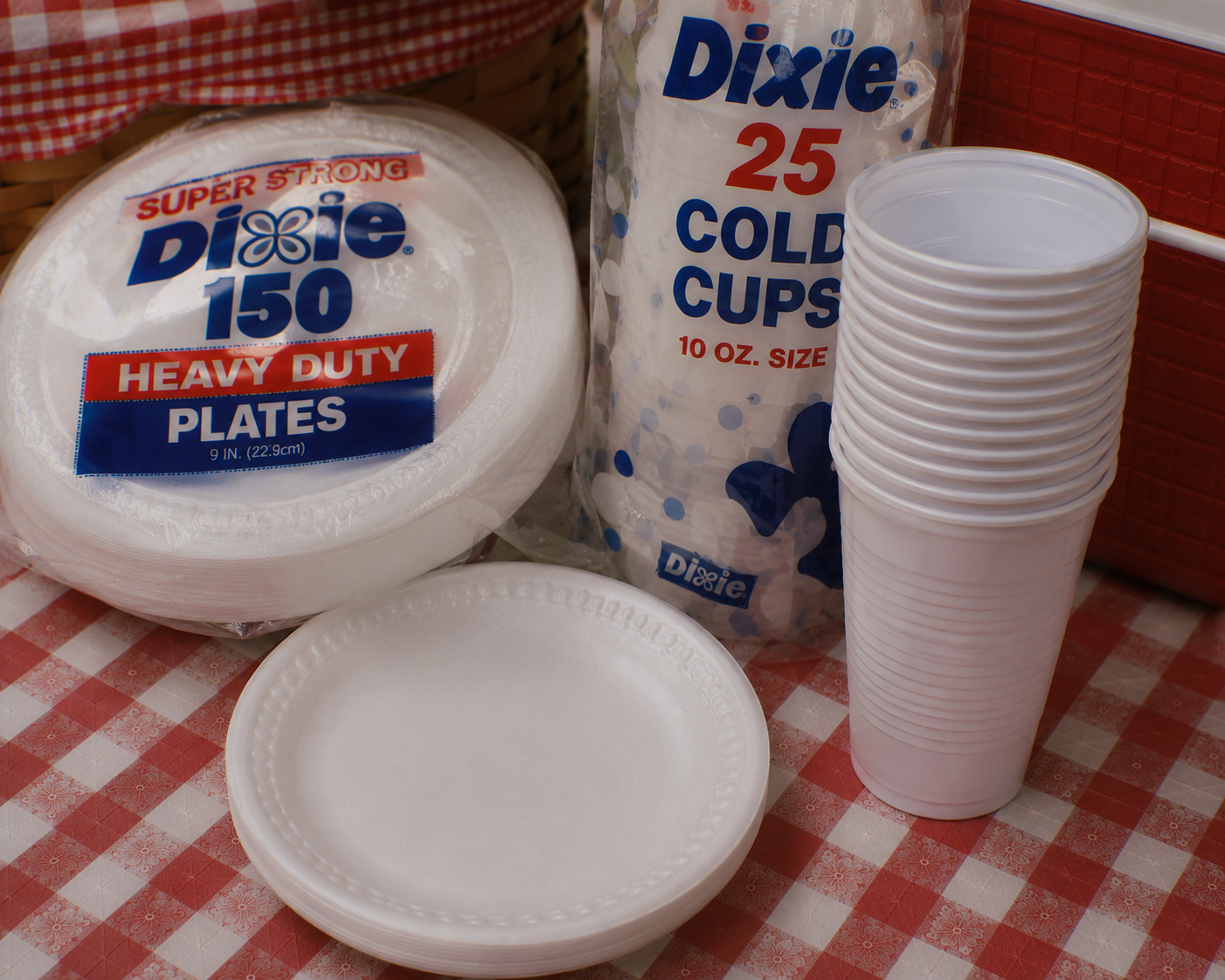 A stack of white foam plates and a sleeve of Dixie cold cups sit on a red and white checkered tablecloth. A few loose plates are stacked in front of the packaging. A red cooler and part of a picnic basket are visible in the background.