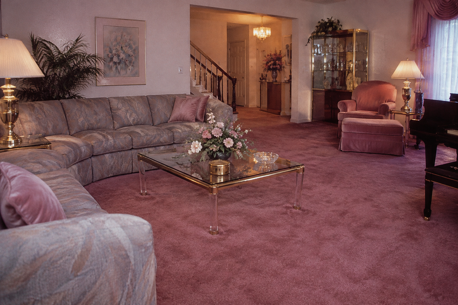 A spacious living room with mauve carpet, a sectional sofa, glass coffee table with flowers, armchair, lamps, a cabinet, and staircase in the background. Soft lighting creates a cozy, elegant atmosphere.