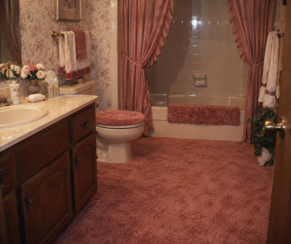 A bathroom with pink carpet, a pink toilet seat cover, pink rugs, floral wallpaper, pink curtains, and a wooden vanity with a sink and flowers.