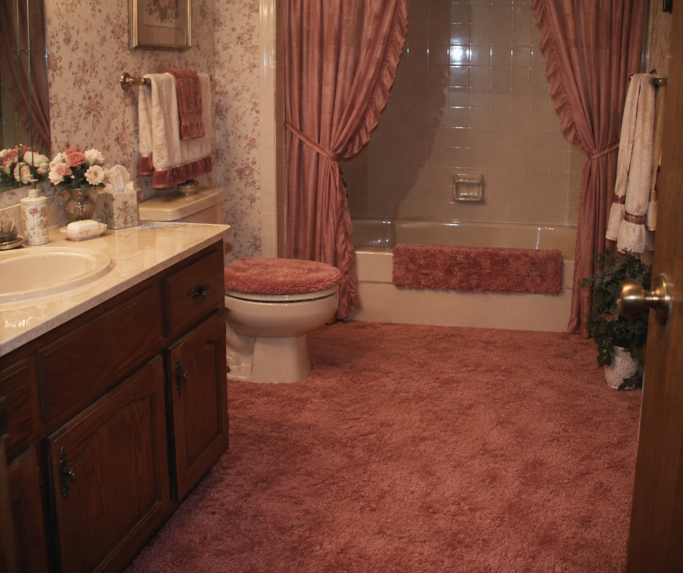 A bathroom with pink carpet, a pink toilet seat cover, pink rugs, floral wallpaper, pink curtains, and a wooden vanity with a sink and flowers.