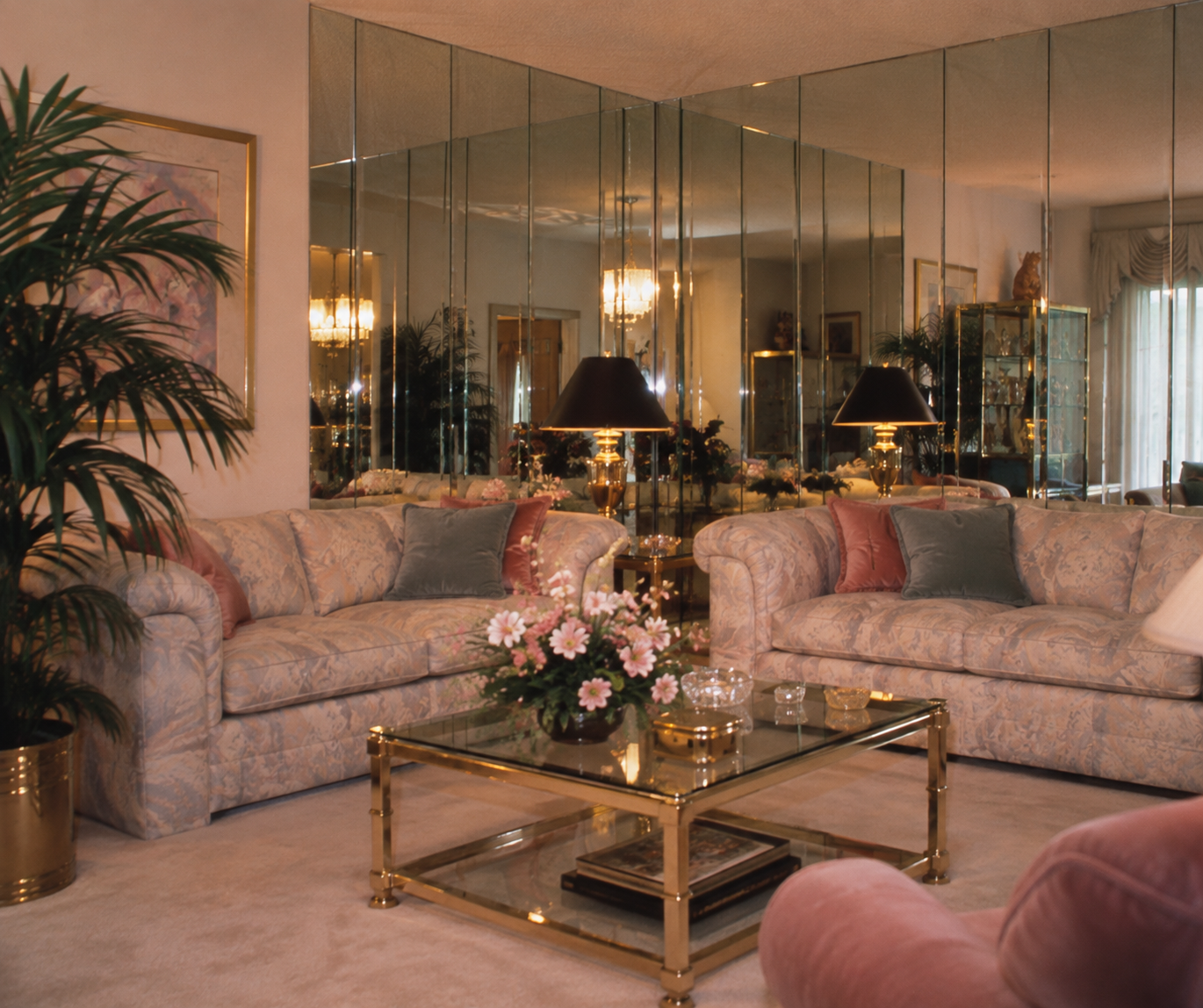A living room with floral sofas, glass coffee table, pink flowers, gold accents, mirrors on the wall, a potted plant, lamps, and soft natural light from a window.