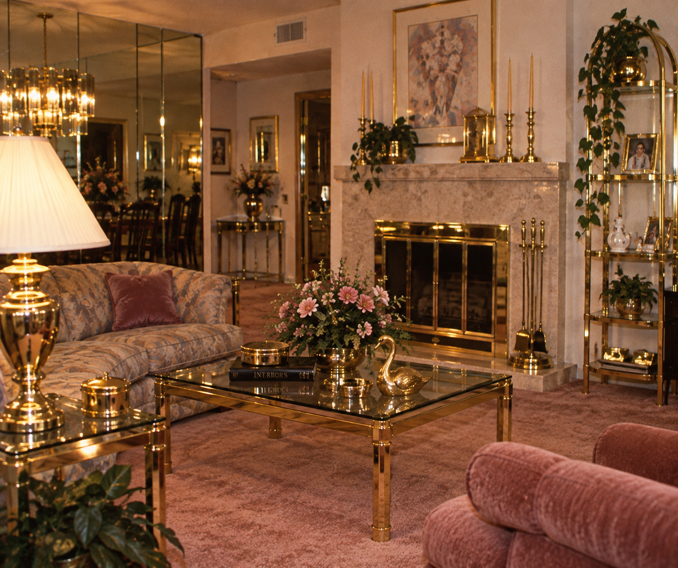 A living room with gold and glass furniture, floral arrangements, pink carpet, a gold-accented fireplace, plush seating, and decorative plants. A dining area is visible in the background through mirrored walls.