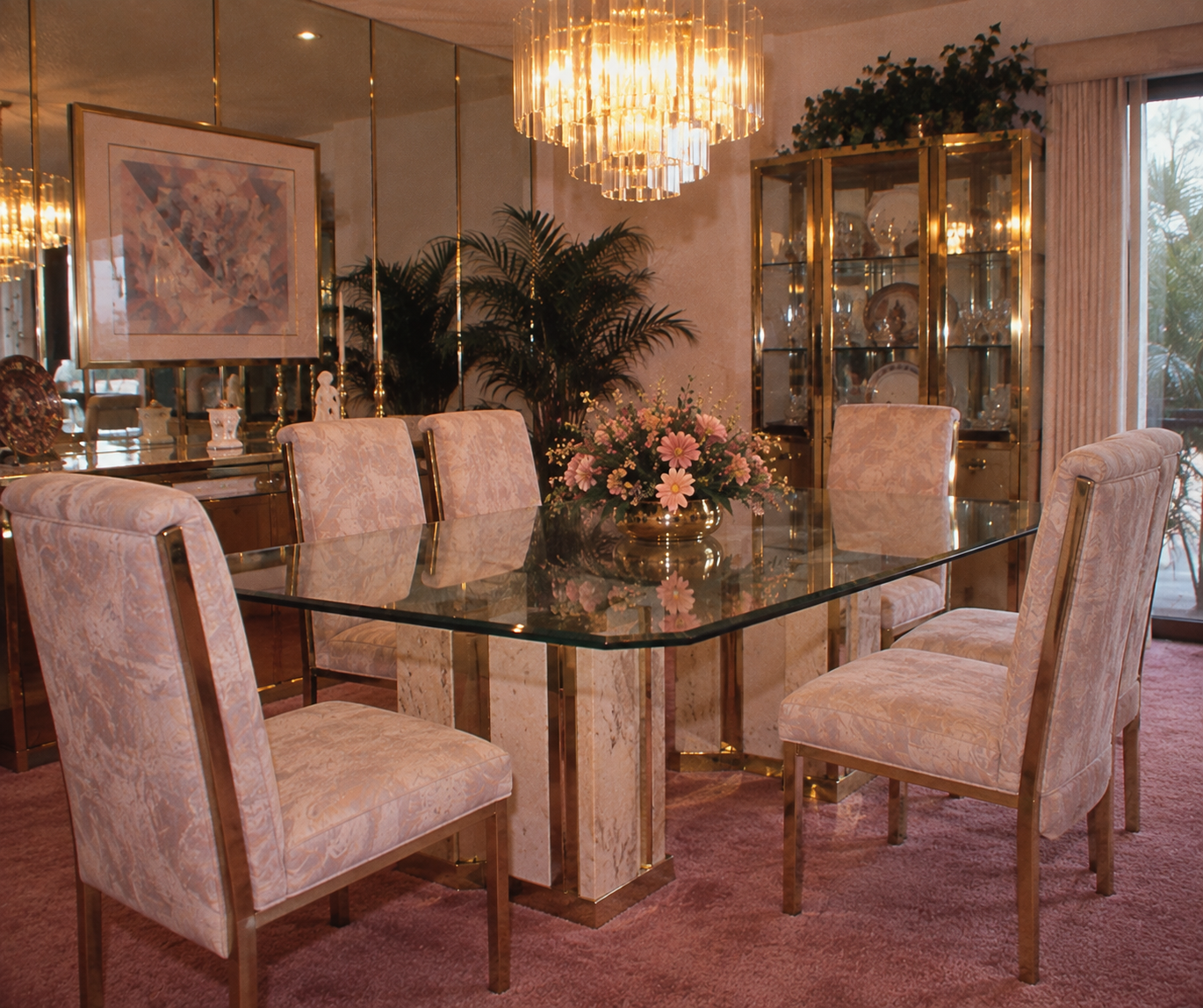 Elegant dining room with a glass table, six upholstered chairs, a floral centerpiece, chandelier, mirrored walls, plants, china cabinet, and pink carpet. Sunlight streams through a sliding glass door.