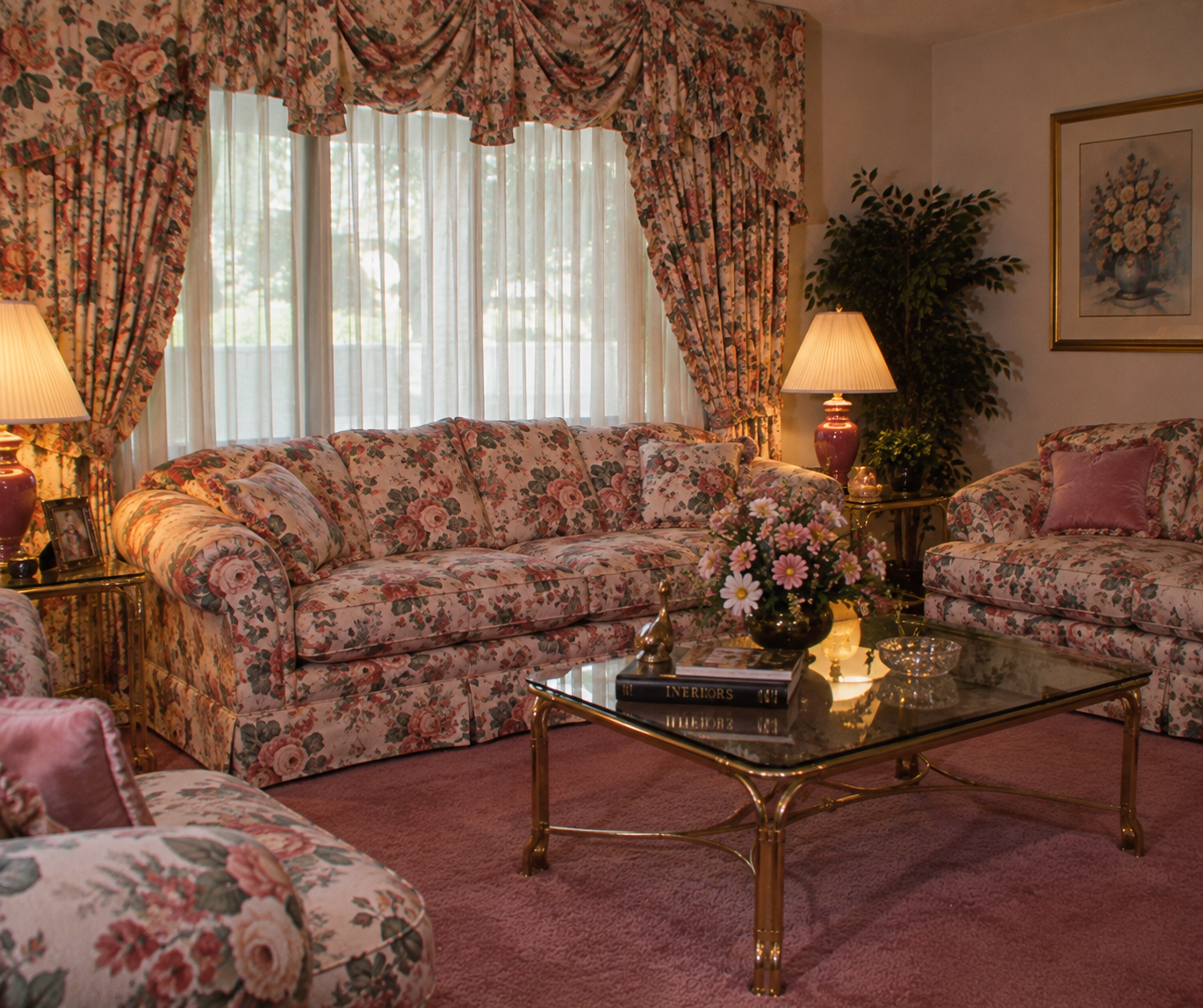 A living room with floral-patterned sofas, matching drapes, two table lamps, a glass coffee table with flowers and books, a framed floral painting, a potted plant, and a pink carpet. Sunlight filters through the window.