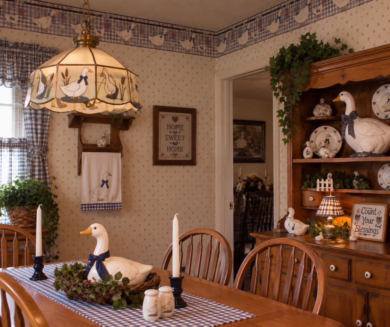 A cozy dining room decorated with a goose theme, featuring goose figurines, a goose-patterned lamp, wooden furniture, candles, plants, and a “Home Sweet Home” sign on the wallpapered wall.