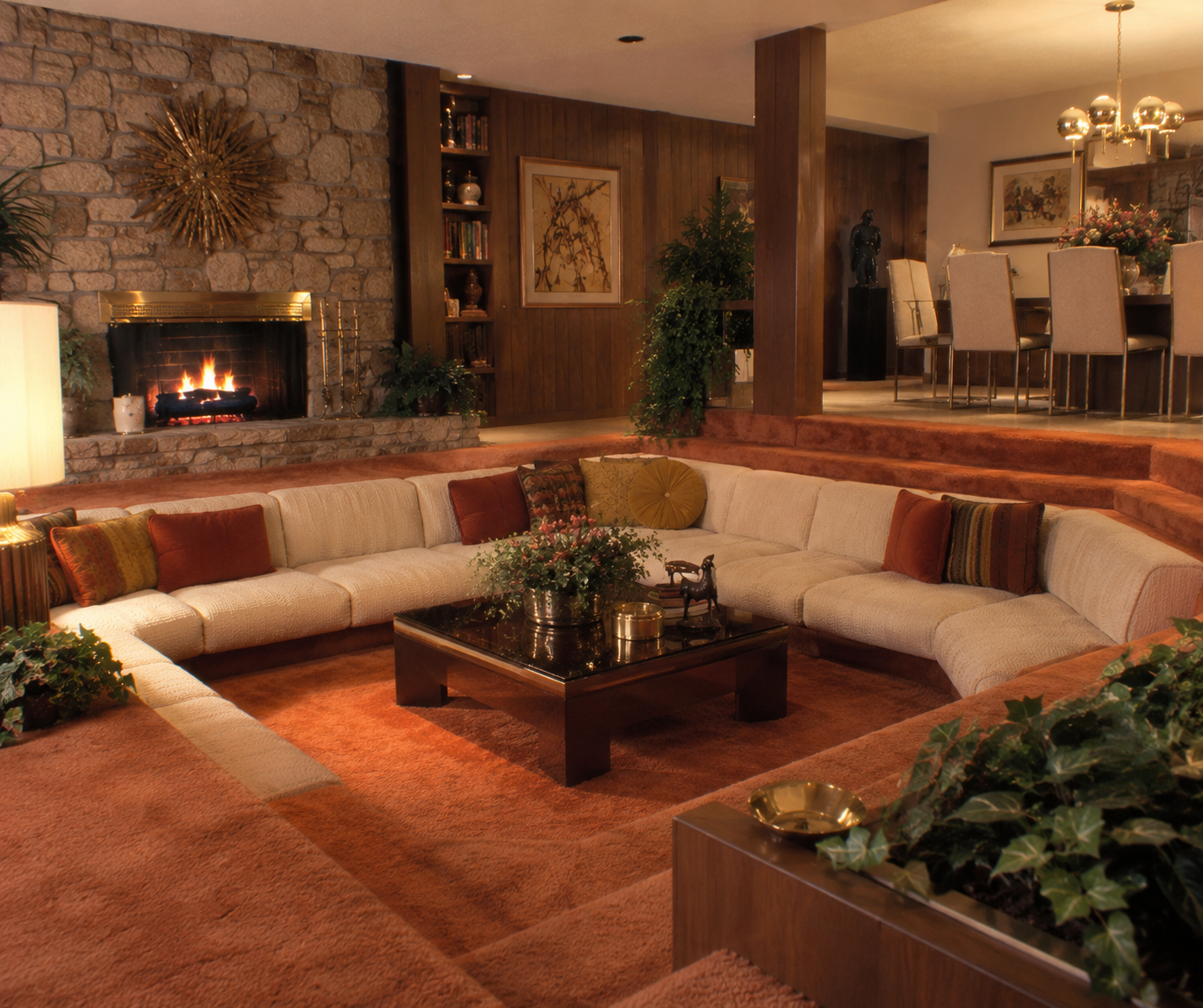 A sunken living room with an L-shaped beige sofa, orange carpet, a dark wood coffee table, plants, a stone fireplace, and a dining area in the background. Warm lighting creates a cozy, retro atmosphere that perfectly captures iconic 1980s home trends.