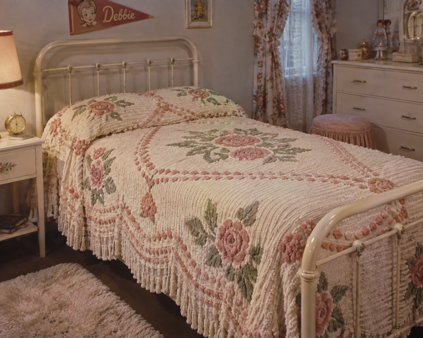 A vintage bedroom with a white metal bed frame, floral chenille bedspread, a nightstand with a lamp, floral curtains, dresser, and a “Debbie” sign on the wall. The room feels cozy and nostalgic.
