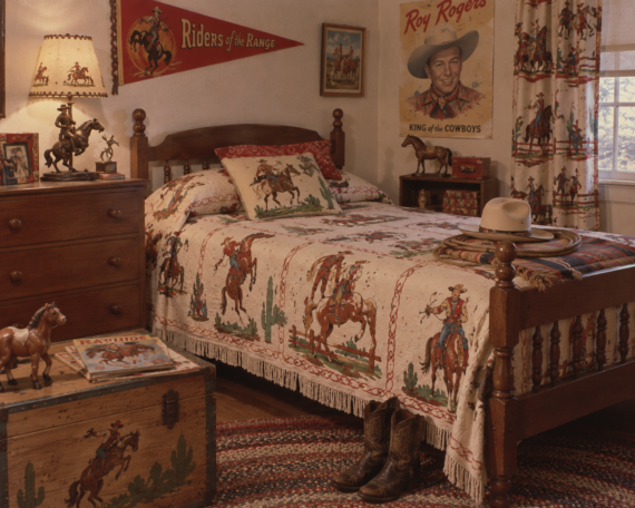 A rustic cowboy-themed bedroom with a wooden bed, western-patterned bedding, cowboy boots, hat, dresser, toys, and Roy Rogers decor. Light filters in through a window with cowboy-print curtains.