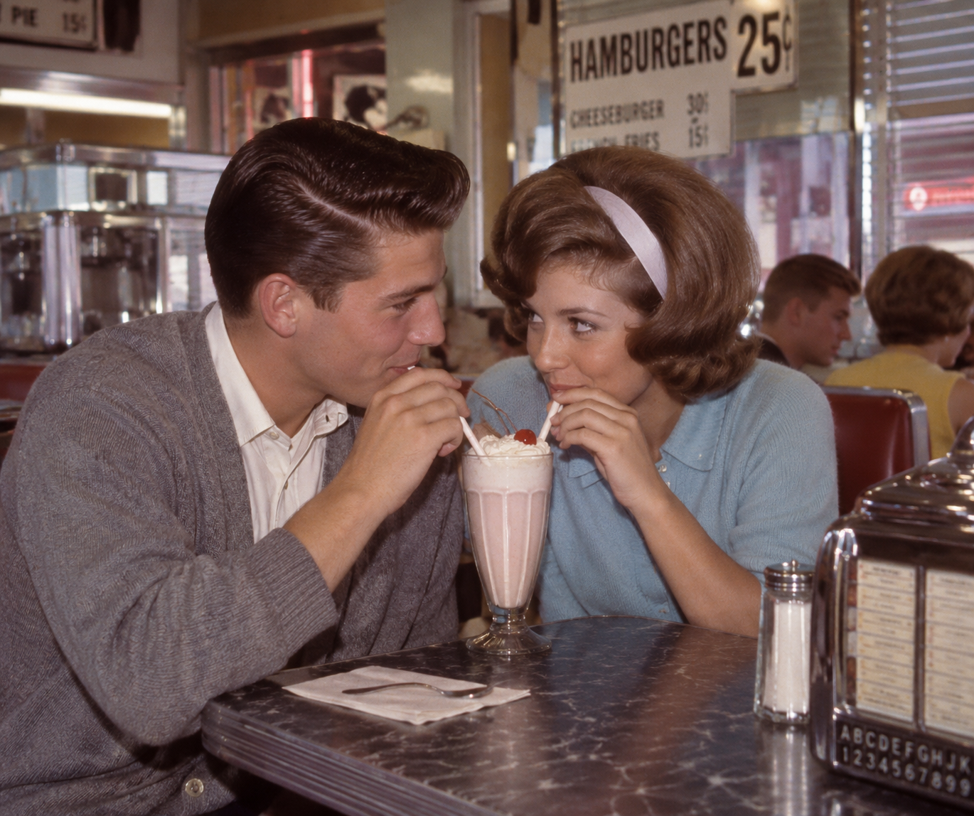 A young couple in 1950s attire shares a pink milkshake at a retro diner, sitting at a table with a jukebox and condiments, smiling at each other.