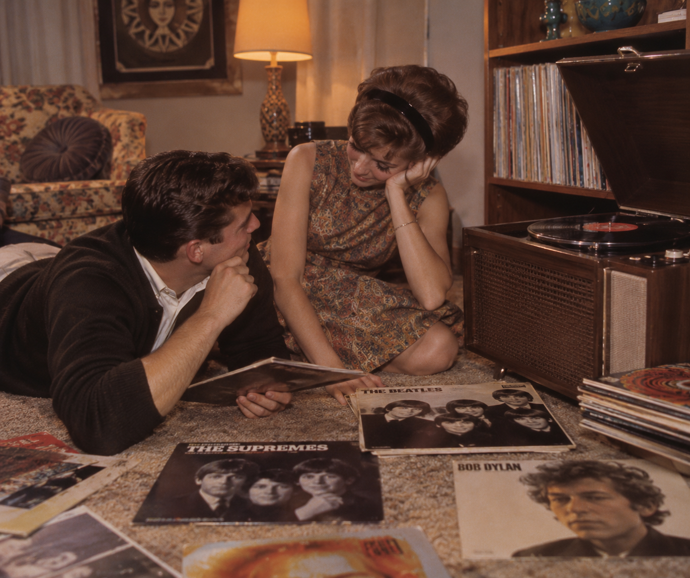 A man and woman sit on the floor by a record player, surrounded by vinyl albums including The Supremes, The Beatles, and Bob Dylan, in a cozy living room with vintage decor.