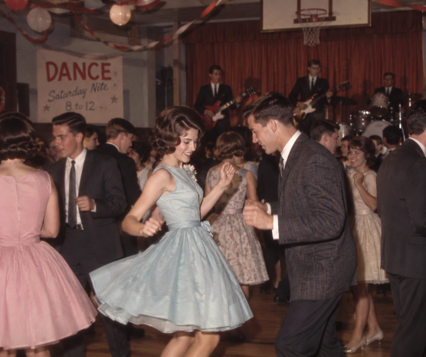 Teenagers in 1960s attire dance energetically in a decorated gym with a live band onstage. A “Dance Saturday Nite” sign and streamers hang in the background. Everyone appears to be having a great time.