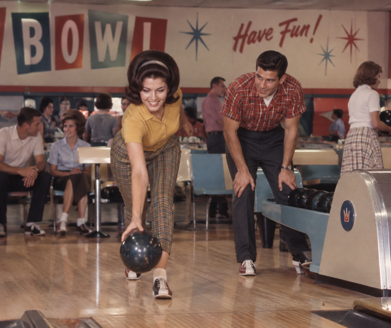A woman in plaid pants bowls while a man in a red plaid shirt watches. Other people sit and chat in the background. The bowling alley is decorated with bright colors and a “Have Fun!” sign.