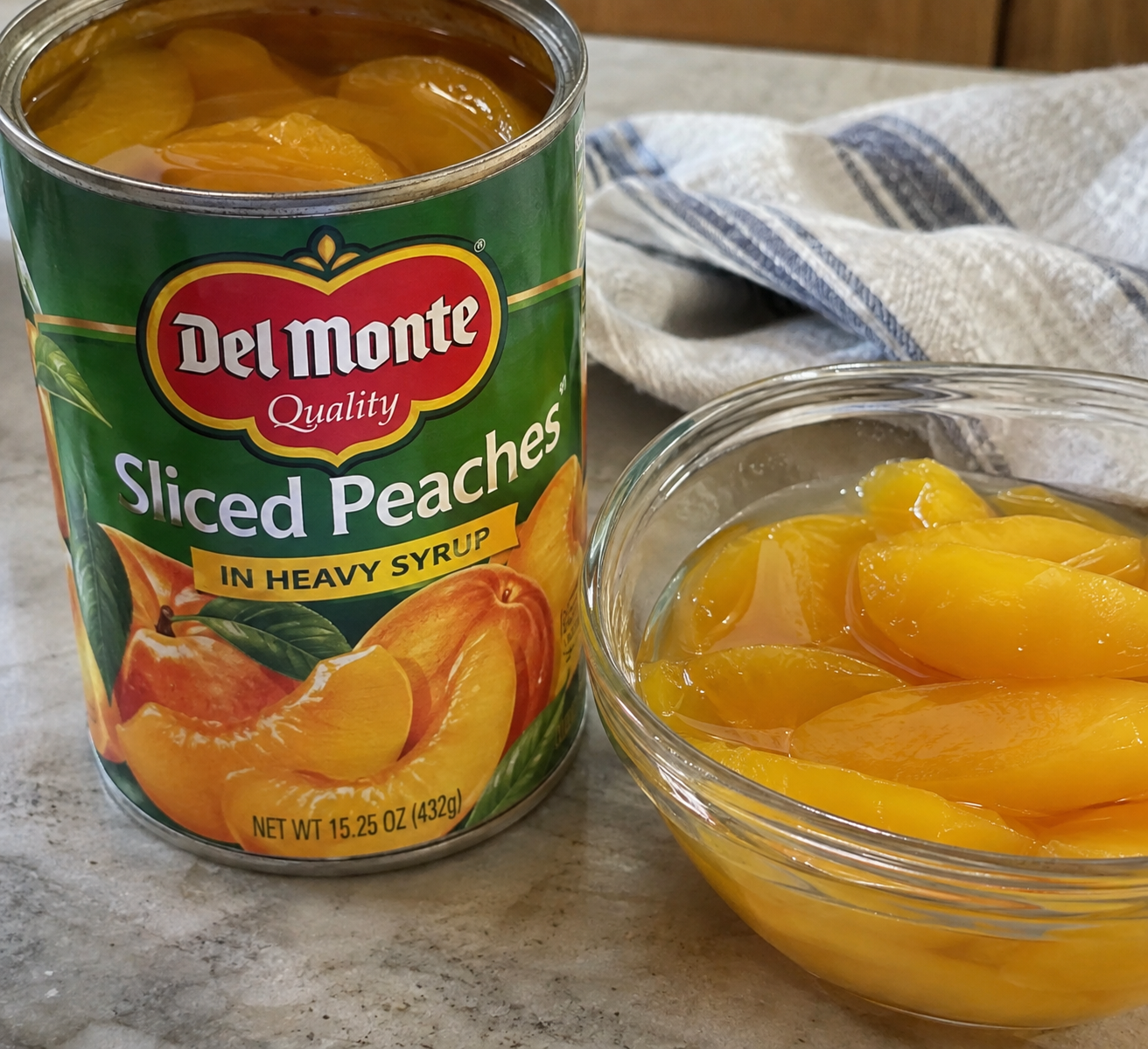 An open can of Del Monte sliced peaches in heavy syrup sits next to a glass bowl filled with the peaches, on a marble countertop with a striped cloth in the background.