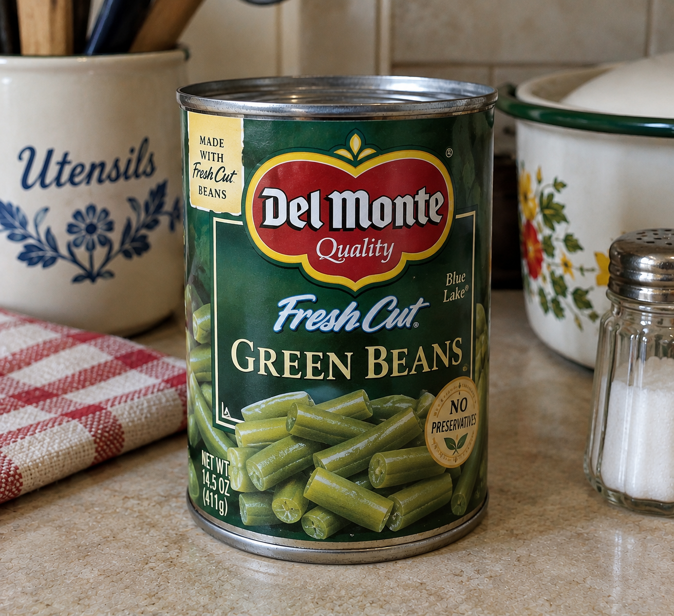 A can of Del Monte Fresh Cut Green Beans sits on a kitchen counter next to a salt shaker, a utensil container, a towel, and a pot with a floral design.