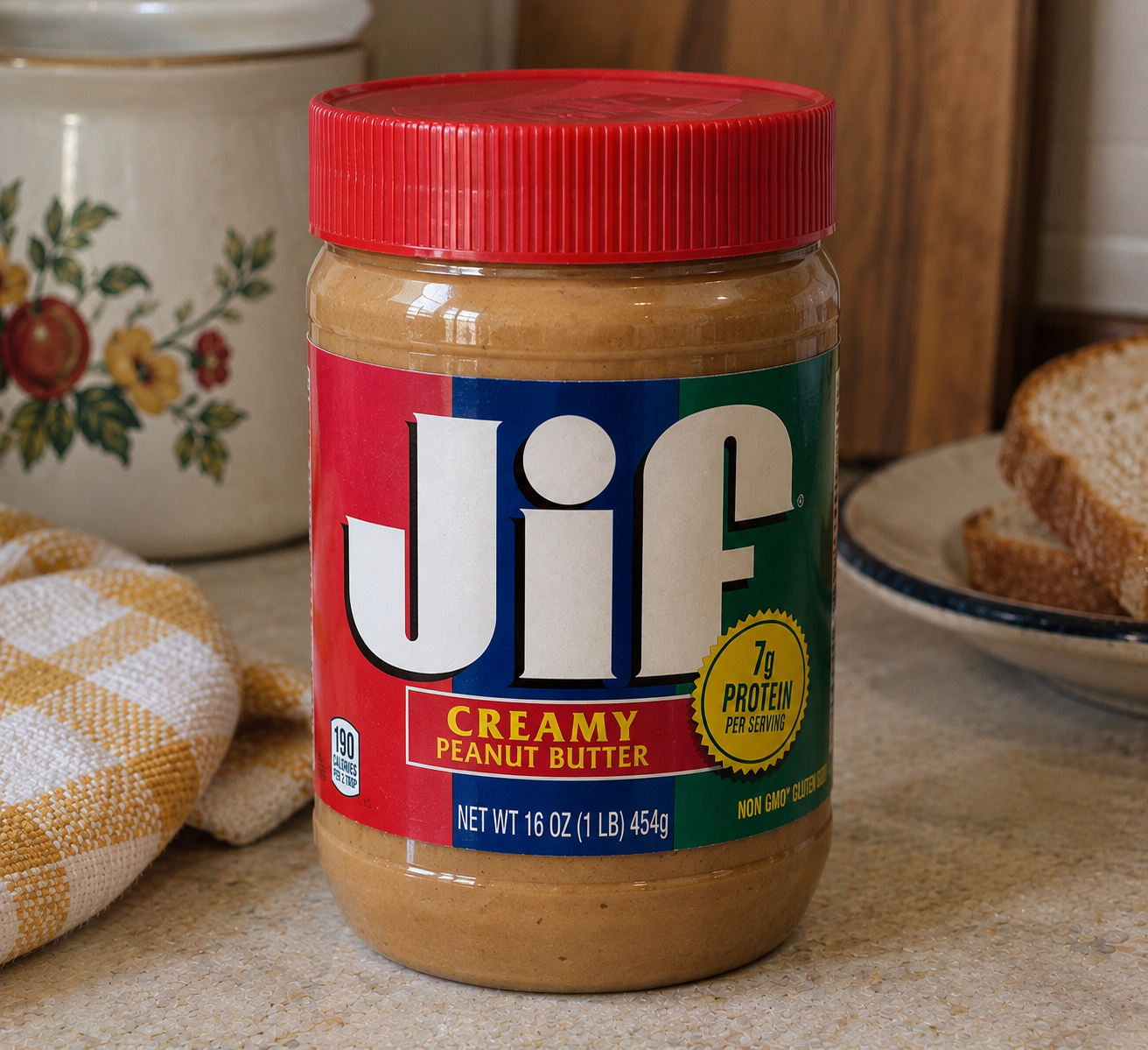A jar of Jif creamy peanut butter sits on a kitchen counter near a plate of sliced bread and a yellow checked towel. The label highlights 7g of protein per serving.