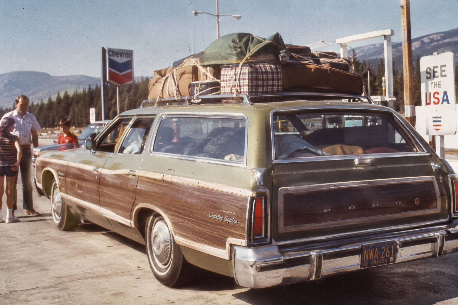 A vintage green station wagon with wood paneling, loaded with luggage on the roof, is parked at a gas station. Several people stand nearby. There’s a Chevron sign and mountains in the background.